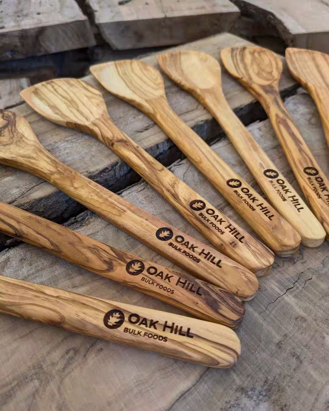 Olive wood spatulas laser-engraved with the Oak Hill Bulk Foods logo and business name, laid out on weathered timber