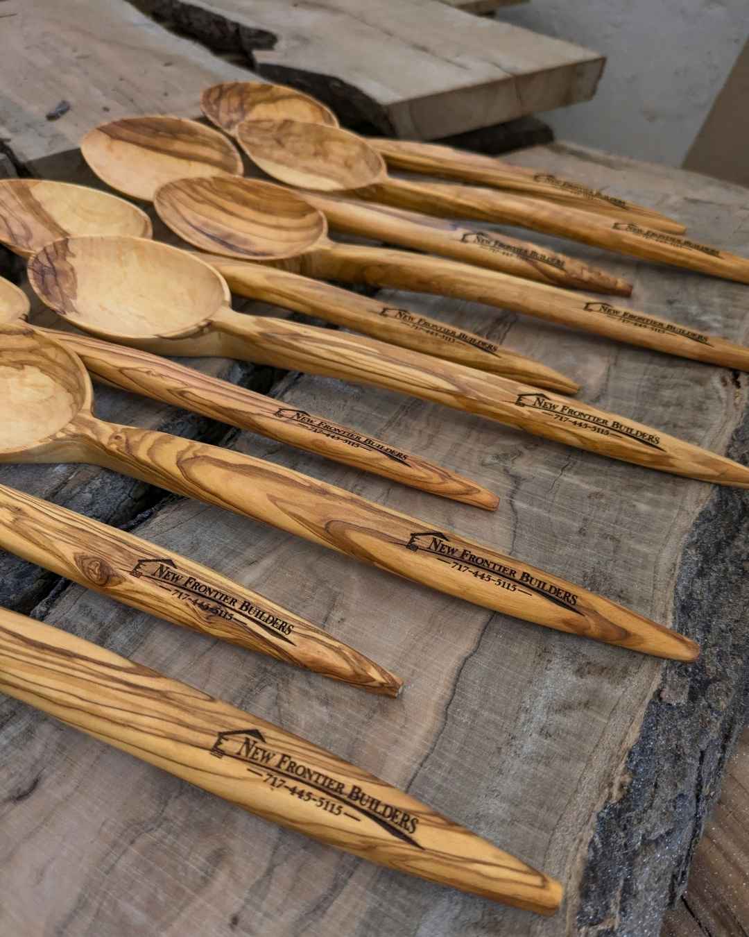 Rows of olive wood spoons laser-engraved with the New Frontier Builders logo and phone number, laid out on rustic wood slabs