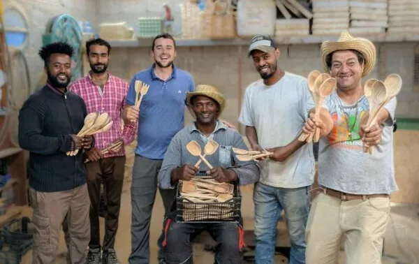 The Lesvos Woodcraft team of refugee artisans standing together in their workshop, holding hand-carved olive wood spoons and utensils