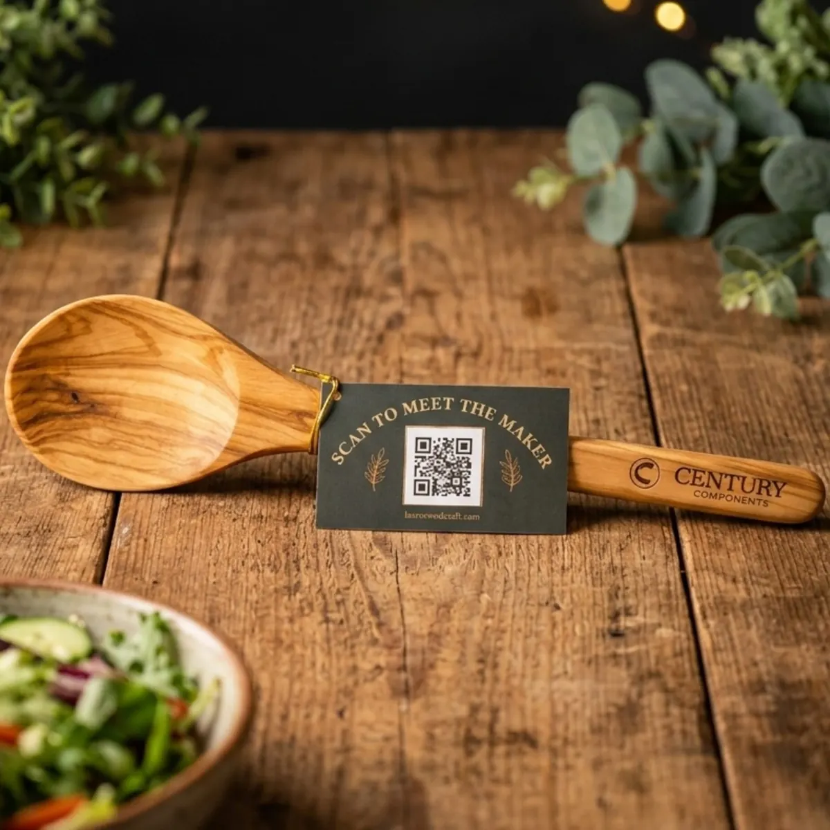 Olive wood serving spoon engraved with the Century Components logo, with a scan-to-meet-the-maker tag on a rustic wood table
