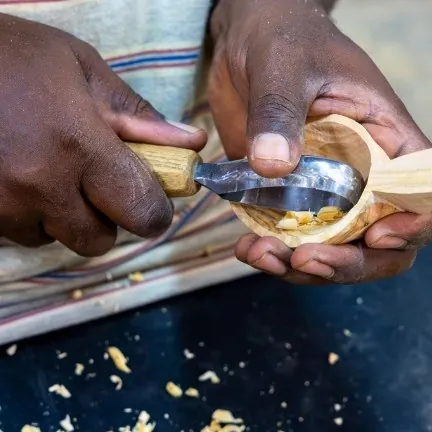 Artisan's hands hand-carving an olive wood spoon with a hook knife, wood shavings on the workbench