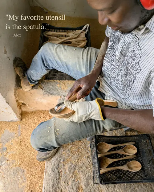 Refugee artisan Alex hand-carving an olive wood spoon, surrounded by finished spoons