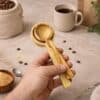 Hand holding a set of handcrafted olive wood coffee scoops in a rustic coffee setting with coffee beans, burlap sack, and coffee mug. Artisan made olive wood scoops from Lesvos Greece with natural grain patterns.